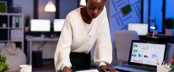 Stressed african manager woman working with financial documents standing at desk checking graphs, holding papers, reading raports late at night in start-up office doing overtime to respect deadline.